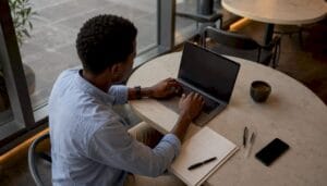 Man with curly hair in a light blue shirt typing on a laptop at a round marble table in a cafe with a notebook pens a smartphone and a cup nearby